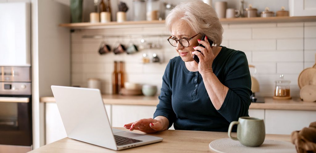 A mature woman double checks she wasn't scammed by calling bank customer service and reviewing her digital banking account on a laptop